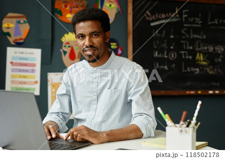Portrait of smiling African American male teacher captured typing on laptop while sitting at working desk in primary school classroom, copy space 118502178