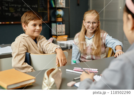 Medium shot of young smiling boy and girl in casual clothing sitting in chairs chatting with friends while enjoying break in classroom at primary school 118502290