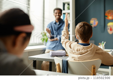Rear view of young male student sitting at desk raising hand to answer teachers question during lesson in primary school classroom, copy space 118502300