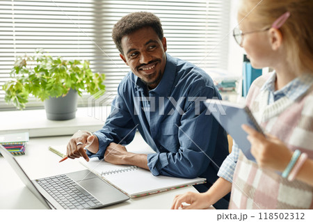 Medium shot of smiling African American male teacher looking at young student while sitting at table with laptop in classroom at primary school, copy space Medium shot of smiling African American male teacher looking at young student while sitting at table with laptop in classroom at primary school, copy space 118502312