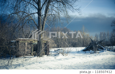 An old abandoned wooden hut sits quietly on a small country road, surrounded by deep winter snow with a dark and gloomy sky above. The crumbling hut is set against a snow-covered field. 118507412
