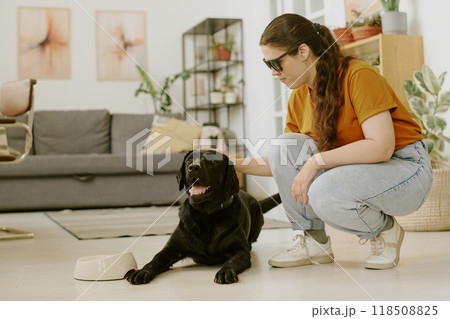 Young woman petting excited black Labrador while he lying on floor Young woman petting excited black Labrador while he lying on floor 118508825
