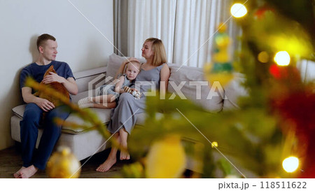 Young loving couple sitting at couch in living room warmth of holiday season playing with baby and cat. Caucasian couple sitting at sofa happily entertaining baby girl and playful cat winter holidays. Young loving couple sitting at couch in living room warmth of holiday season playing with baby and cat. Caucasian couple sitting at sofa happily entertaining baby girl and playful cat winter holidays. 118511622