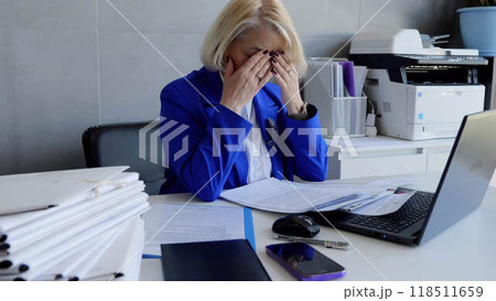 Middle aged businesswoman in suit sits in office holding head overwork exhaustion emotional stress. Sitting in office caucasian businesswoman in suit shows signs of fatigue and holds her head. 118511659