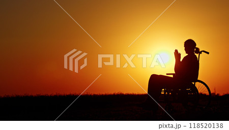 Silhouette of a person in a wheelchair at sunset, hands clasped in quiet reflection. 118520138