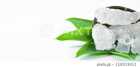 Clear and pristine alum cubes resting in a bamboo basket alongside leaves, set against a white background. 118533011