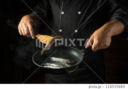 Cooking fish in a frying pan. A cook fries capelin in a restaurant kitchen before dinner. A spatula with a small fish in the chef hand Cooking fish in a frying pan. A cook fries capelin in a restaurant kitchen before dinner. A spatula with a small fish in the chef hand 118535669
