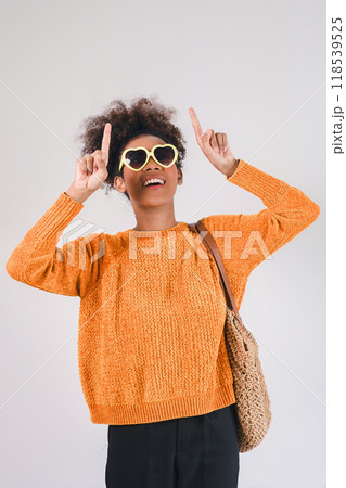 Stylish african american woman in orange sweater wearing heart-shaped sunglasses, pointing upward. Isolated on white background Stylish african american woman in orange sweater wearing heart-shaped sunglasses, pointing upward. Isolated on white background 118539525