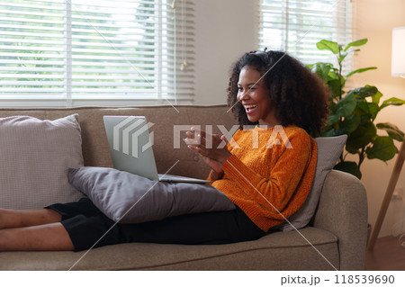 Smiling African American woman enjoying online content on laptop while relaxing on comfortable couch at home. Smiling African American woman enjoying online content on laptop while relaxing on comfortable couch at home. 118539690