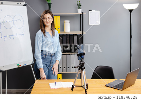 Positive blogger woman speaking at smartphone webcam on tripod, recording post for vlog. Online teacher, coach working, giving webinar at home. Presenter taking video on cellphone 118546328