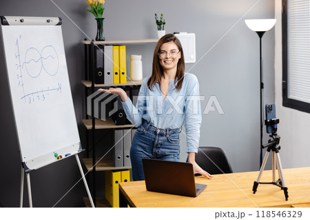 Videoconference And Training. Class of diverse students learning remotely online using web app. Female teacher providing lesson with laptop, recording video on cellphone on tripod 118546329