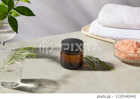 Spa setting photo featuring an unbranded amber cosmetic pot on a gray table top, next to a glass bowl of himalayan pink salt and white towels rolled up, which placed on beige tray. High angle view Spa setting photo featuring an unbranded amber cosmetic pot on a gray table top, next to a glass bowl of himalayan pink salt and white towels rolled up, which placed on beige tray. High angle view 118546942
