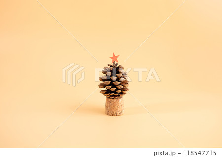 Pine cone on cork base with red star topper, light orange background, simple and sustainable Christmas tree concept, natural holiday decoration, minimalist design, alternative festive decor, 118547715