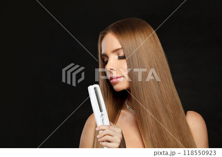 Stylish woman with hair straightener iron in studio isolated on black background. Haircare, wellness 118550923