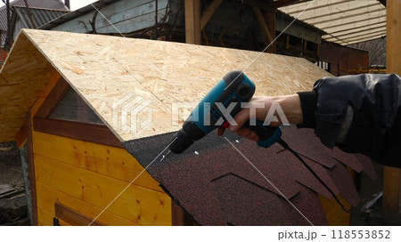 attaching roofing felt to a wooden roof of a chicken coop using a screwdriver, building a wooden structure for poultry, finishing the roof with a soft covering, tightening the fasteners 118553852