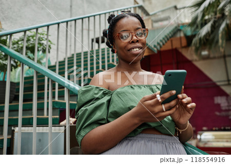 Portrait of young African American woman holding smartphone and looking at camera Portrait of young African American woman holding smartphone and looking at camera 118554916