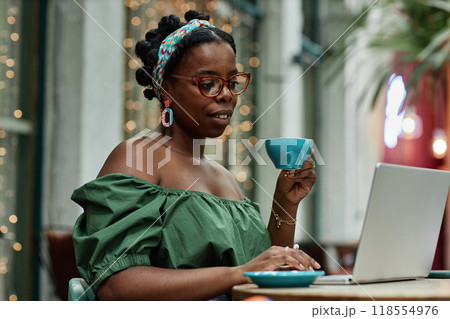 Young African American woman holding cup of tea while surfing in Internet in cafe Young African American woman holding cup of tea while surfing in Internet in cafe 118554976