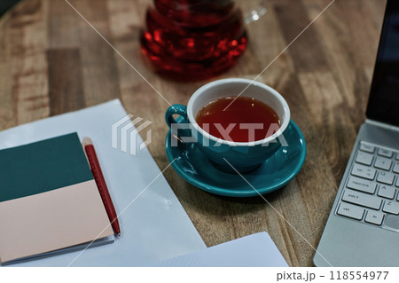 High angle shot of turquoise ceramic cup of black tea on wooden table with laptop and notes High angle shot of turquoise ceramic cup of black tea on wooden table with laptop and notes 118554977