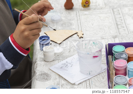 Child hands with a brush and paints. Boy colorize a wooden figurine on the table, selective focus. 118557547