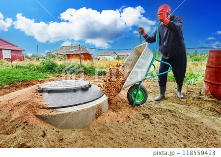 Worker buries concrete rings of septic tank with sand, pouring it out of wheelbarrow. Worker buries concrete rings of septic tank with sand, pouring it out of wheelbarrow. 118559413