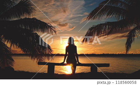 Silhouette of a woman sitting on a bench by the sea, enjoying a beautiful sunset over a tropical island with palm trees Silhouette of a woman sitting on a bench by the sea, enjoying a beautiful sunset over a tropical island with palm trees 118560058