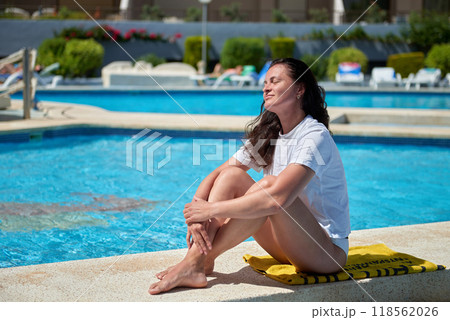 Serene Summer Escape: Young Woman Relaxing Poolside in Elegant Pose, Soaking Up Sun at Luxurious Resort Getaway. Blissful Poolside Retreat: Carefree Female Sunbathing on Edge of Infinity Pool 118562026