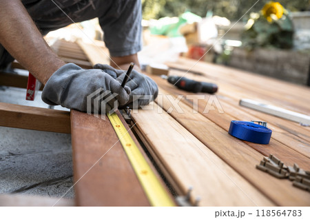 Worker with protective gloves marking a spot on wooden plank while placing it on a foundation for outside patio 118564783