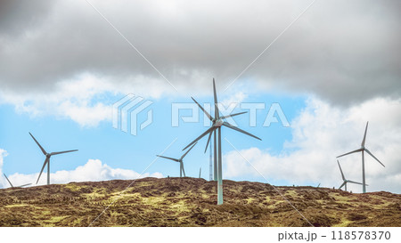 Few wind turbines stand on a grassy hill against a backdrop of blue sky and white clouds. 118578370