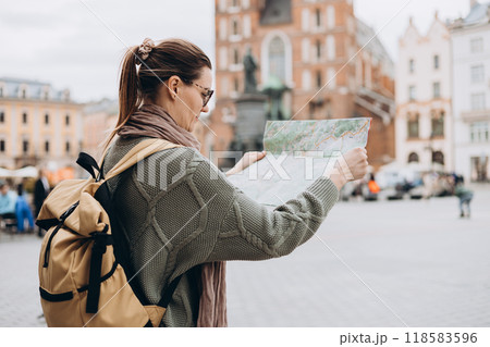 Attractive young female tourist is exploring new city. Happy girl holding a paper map on Market Square in Krakow. Traveling Europe in autumn. St. Marys Basilica Attractive young female tourist is exploring new city. Happy girl holding a paper map on Market Square in Krakow. Traveling Europe in autumn. St. Marys Basilica 118583596