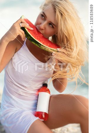 a beautiful blonde girl outdoors eats a watermelon with emotions and a bottle of red drink with her a beautiful blonde girl outdoors eats a watermelon with emotions and a bottle of red drink with her 118583689