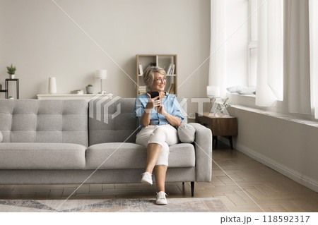 Mature woman sits on sofa with smartphone in living room 118592317