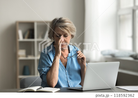Woman taking off her eyeglasses seated at desk with laptop Woman taking off her eyeglasses seated at desk with laptop 118592362