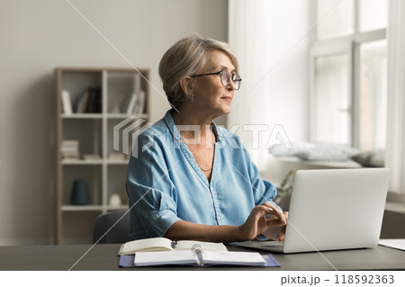 Woman sits at desk with laptop, looking thoughtfully out window 118592363