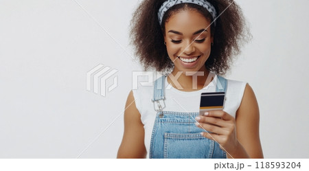 Smiling woman in overalls holding a phone and credit card, expressing happiness and confidence in online shopping 118593204