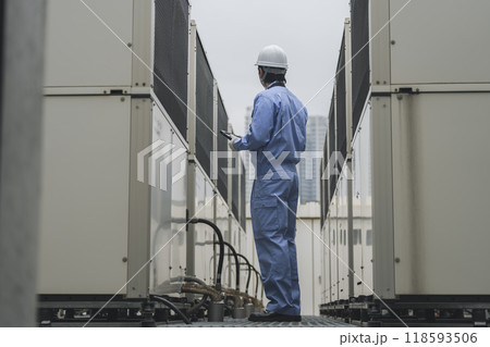 ビルメンテナンス　イメージ　ビル屋上に設置された空調の室外機を点検する作業服姿の男性 118593506