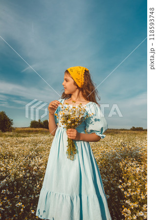 Happy natural girl in blue linen dress and yellow knitted kerchief stands and holds bouquet of flowers in daisy field 118593748