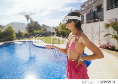 A woman wearing a virtual reality headset while relaxing by the poolside in a sunny paradise A woman wearing a virtual reality headset while relaxing by the poolside in a sunny paradise 118593914