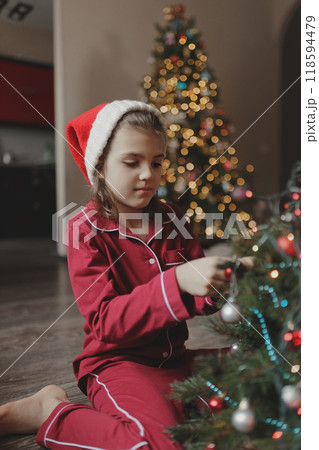 Portrait of girl of 7 years old in red pajamas and Santa Claus hat sitting on living room floor decorates Christmas tree Portrait of girl of 7 years old in red pajamas and Santa Claus hat sitting on living room floor decorates Christmas tree 118594479