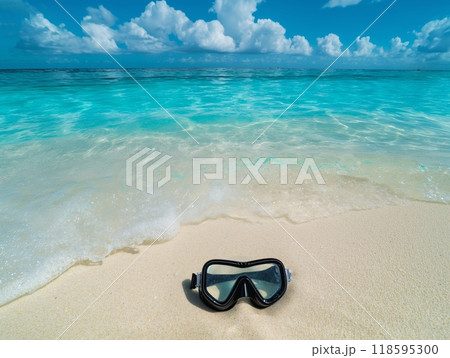 A black snorkel mask lies on a sandy beach with clear turquoise waters and a blue sky with fluffy clouds. 118595300
