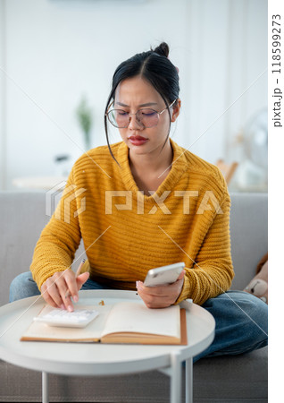 A serious, thoughtful Asian woman sits on a couch in the living room, worriedly planning her budget. 118599273