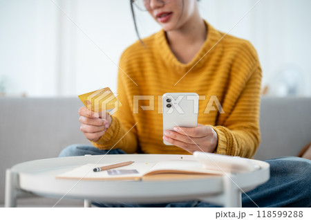 A woman sitting on a couch and holding her smartphone and a credit card, shopping online at home. A woman sitting on a couch and holding her smartphone and a credit card, shopping online at home. 118599288