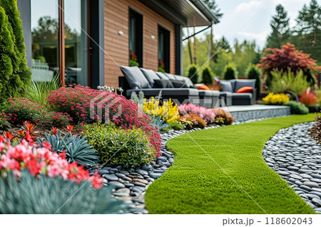 Garden with a stone path and a green lawn. Full of colorful flowers and plants, and there are several chairs and couches placed around the area for relaxation 118602043