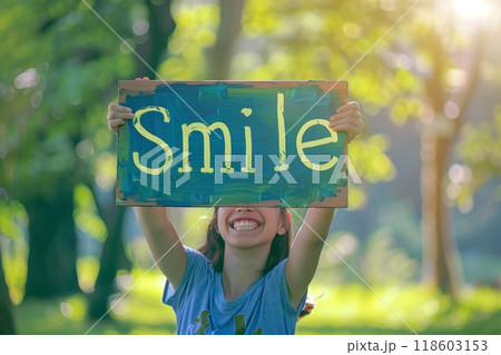 A person holds up a cheerful hand-painted sign with the word Smile in a lush green park, spreading positivity and optimism in a peaceful natural setting 118603153