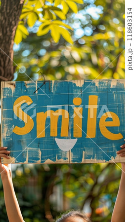 In a vibrant park filled with greenery, an individual shares an uplifting message of happiness by holding a colorful sign that says Smile, symbolizing optimism and joy 118603154