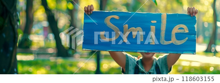A hand-painted sign with the word Smile is held by a person in a lush park, promoting a simple yet powerful message of positivity, hope, and connection with nature, banner 118603155