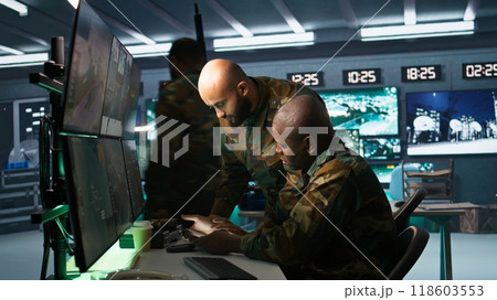 Military dispatcher helped by coworker with tablet to gain info from army intelligence analysts. Army intelligence corps expert discussing with colleague in command post, camera B 118603553