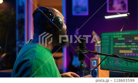 Confident artist singer performing a song on microphone in home studio, recording his audio files and manipulating raw sound on professional editing software. Music production. Camera A. Confident artist singer performing a song on microphone in home studio, recording his audio files and manipulating raw sound on professional editing software. Music production. Camera A. 118603599
