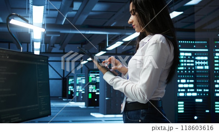Data center computer scientist designing, implementing and managing infrastructure to ensure optimal performance and reliability. Woman in server room coding on tablet, camera B Data center computer scientist designing, implementing and managing infrastructure to ensure optimal performance and reliability. Woman in server room coding on tablet, camera B 118603616
