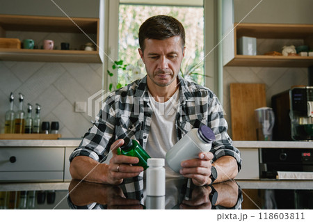 Man is sitting at table in kitchen looking at various bottles of medicine capsules. Person is taking pills or vitamins. Concept of supplements, medicine or health care 118603811