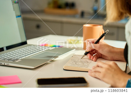 Person is sketching in notebook while working remotely at desk with laptop. Freelance work at home office. Graphic designer drawing sketch Person is sketching in notebook while working remotely at desk with laptop. Freelance work at home office. Graphic designer drawing sketch 118603841